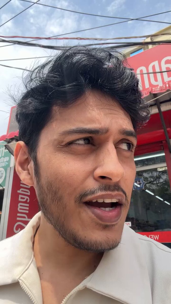 The image features a man wearing a white shirt who appears to be making an odd face or expression as he looks off to the side, possibly reacting to something that has happened or someone saying something. The photo was taken by another person, likely a friend of his, and it is being shared on social media platforms. The post includes text such as "damn people are actually using @KASTxyz card for irl shopping in Bangladesh now ngl i never expected to see this working in bd this early the way crypto moving now with @BitgetWallet & now kast in bd, feeling something good on way thoon video by hisham iqbal -top anime cc".