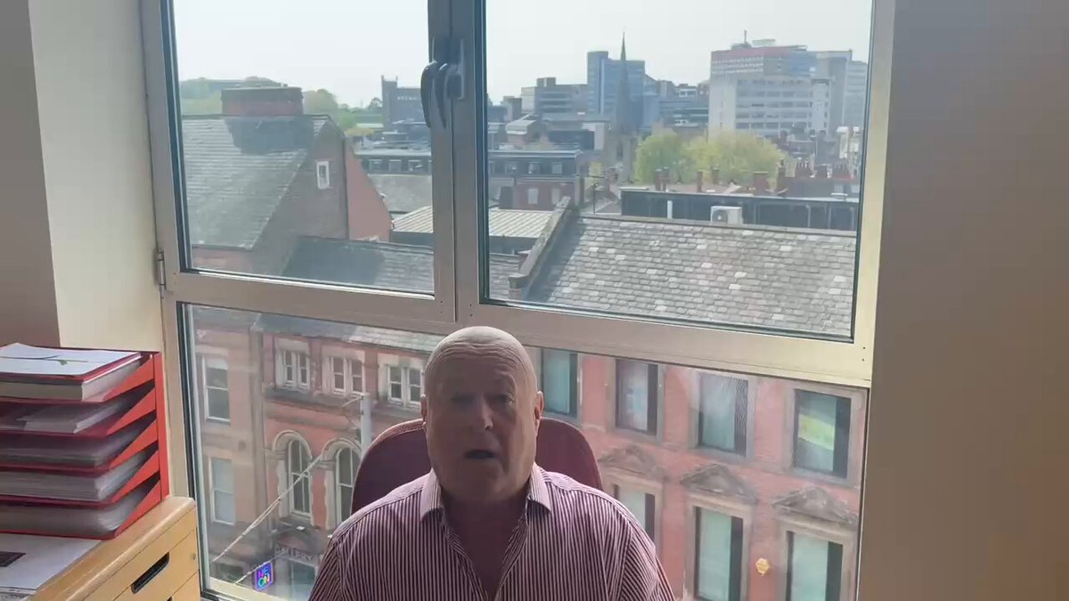 The image shows a man sitting at his desk with a window behind him, looking out over a cityscape. The man appears to be wearing a striped shirt and has a serious expression as he looks out the window. There are several books scattered around the room, suggesting that this might be an office or workspace setting.