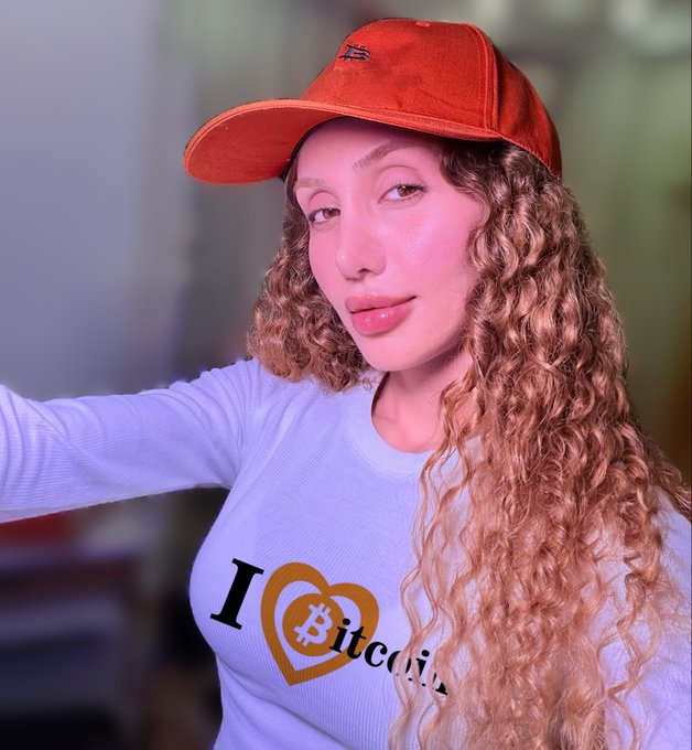 A young woman with long, curly reddish-blonde hair wearing a red baseball cap and a white long-sleeve shirt that says 'I ❤️ Bitcoin', appearing to take a selfie.