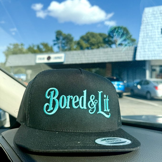 A black snapback hat with teal embroidered lettering reading 'Bored & Lit' and a 'Classic Snapback' sticker on the brim rests on a car dashboard, with a building visible through the windshield.