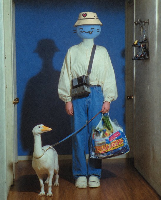 A person wearing a blue cartoon character head mask, a beige jacket, and blue jeans stands in a doorway holding a leash attached to a white duck, along with a plastic shopping bag and a Game Boy camera.