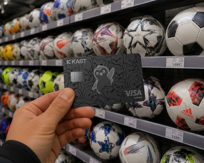 A hand holds a black KAST Visa Infinite credit card with a ghost design in front of a store shelf displaying various Adidas soccer balls priced at $1,699. The image illustrates KAST's crypto-based payment solution by placing the card in a relatable retail setting, emphasizing the company's focus on solving everyday financial tasks like payments.