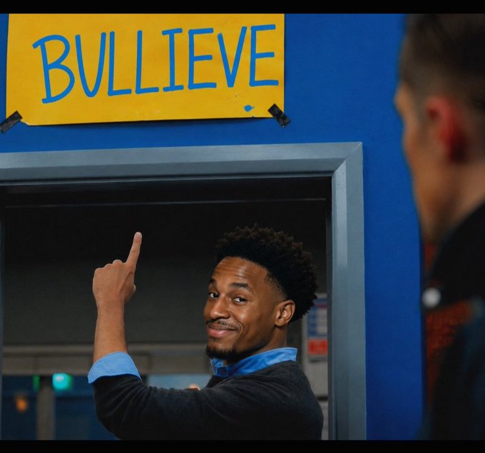 A young man in a blue shirt points upward at a yellow sign reading 'BULLIEVE' taped to a blue wall, while another person is partially visible in the foreground.