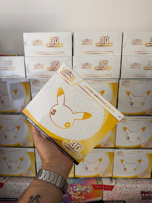 A hand holds a Pokémon TCG 'Coffret Dresseur d'Élite' 30th Anniversary Elite Trainer Box featuring Pikachu, with multiple boxes stacked in the background.