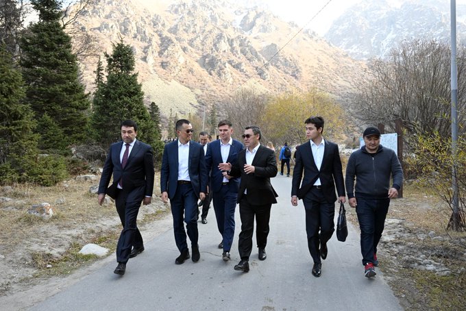 A group of men in suits walk along a paved road with rocky mountains and evergreen trees in the background.