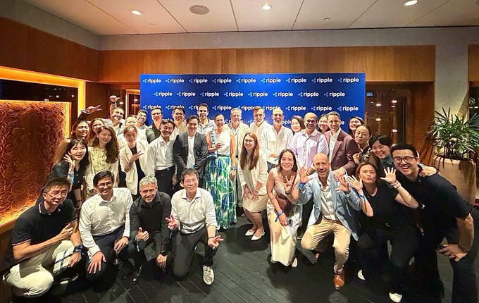 A large group poses in front of a blue backdrop displaying the "ripple" logo in a repeating pattern, gathered for a corporate event. The celebratory photo captures Ripple's APAC team and clients together, visually supporting the announcement of their major client celebration in Singapore and their expansion into the Australian market.