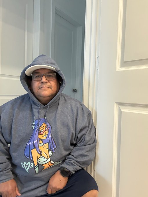 A man wearing glasses and a grey hoodie featuring a cartoon anime-style character design sits on a chair in front of white doors.