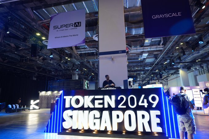 A conference venue with a large illuminated "TOKEN 2049 SINGAPORE" sign in the foreground and hanging banners for "SUPER AI" and "GRAYSCALE" in the background.