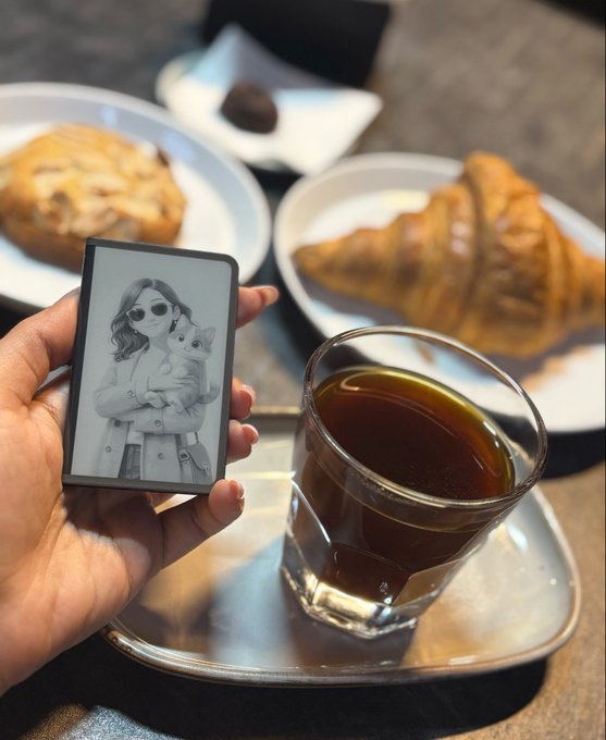 A hand holding a small digital photo frame displaying a black-and-white image of a woman with sunglasses holding a cat, alongside a coffee, croissant, and pastry on a café table.