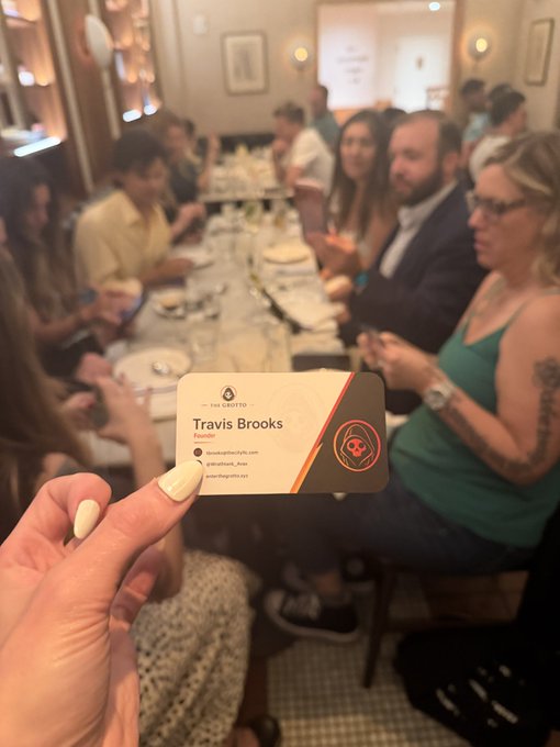 A hand holds a business card for 'Travis Brooks, Founder' with contact information, while a group of people sit around a dining table in the blurred background.