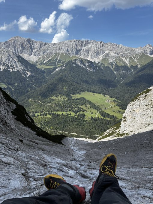 A first-person view looking down at hiking boots and legs dangling over the edge of a rocky mountain cliff, overlooking a green alpine valley with a small town and dramatic mountain peaks under a blue sky.