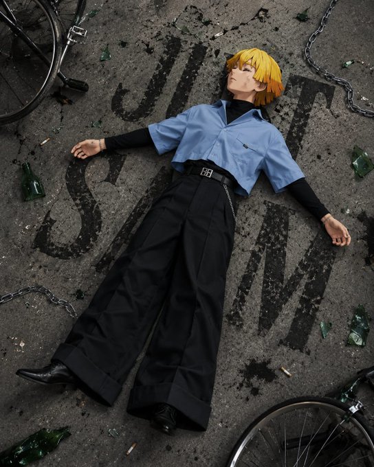 A cosplay photo of a person with blonde hair in a blue shirt and black pants lying on the ground surrounded by broken glass, bicycle wheels, and chains.