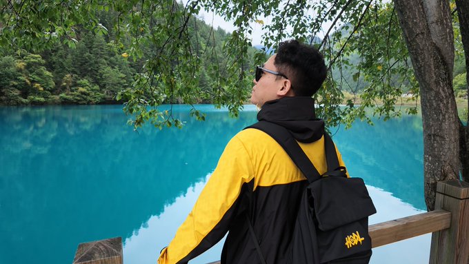 A person wearing a yellow and black jacket with a backpack stands by a wooden railing, looking out at a striking turquoise lake surrounded by forest.