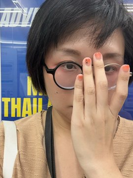 A selfie of a woman with short dark hair and round glasses, partially covering her face with her hand showing red-painted nails.