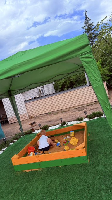 A young child plays in a wooden sandbox filled with toys under a green canopy tent set up on artificial turf in a backyard.