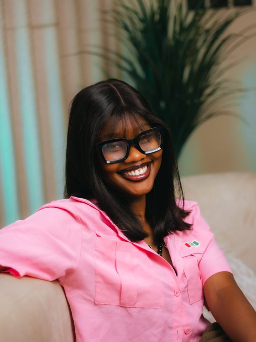 A smiling woman wearing black-rimmed glasses and a bright pink button-up shirt sits on a beige couch with a green plant and curtains visible in the background.