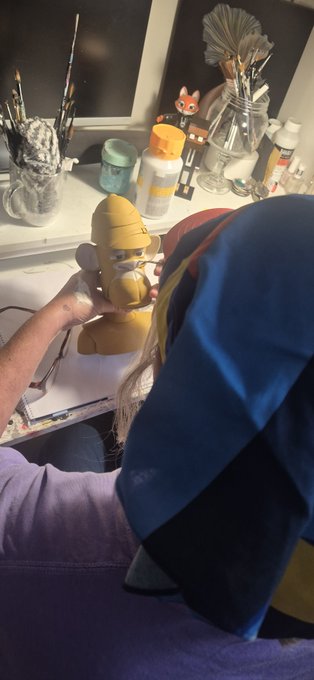 An artist is working on a yellow resin sculpture at a workspace filled with paintbrushes, bottles, and a small fox figurine, with various tools scattered around.