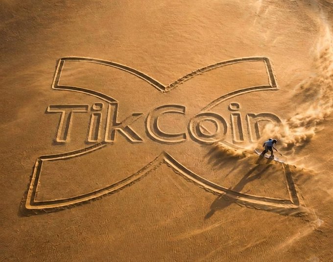 Aerial photograph showing the TikCoin logo etched into sand, with a person walking across the design kicking up dust.