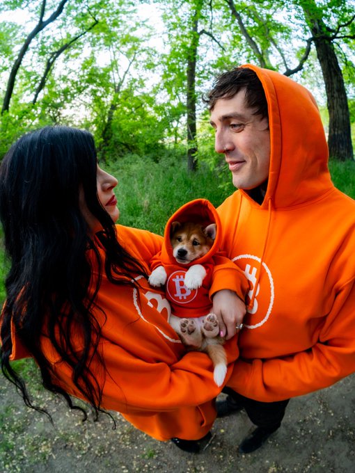 A couple in matching orange Bitcoin-themed hoodies hold a corgi puppy also dressed in an orange Bitcoin hoodie, standing in a lush green outdoor setting.