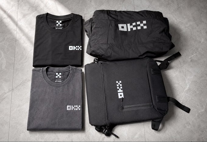 An overhead product shot displaying OKX-branded merchandise: two folded t-shirts (black and gray), a black jacket, and a black backpack arranged on a gray surface.
