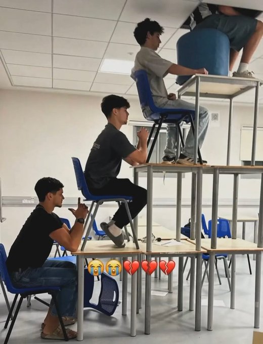 A classroom photo showing several students seated on chairs stacked on top of desks at progressively higher levels, with crying and red heart emojis overlaid at the bottom.