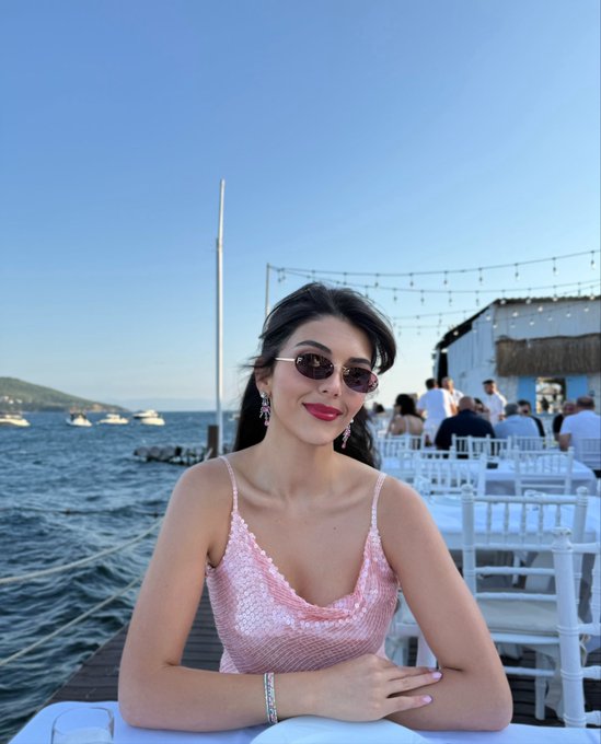 A woman with dark hair, red lipstick, and sunglasses wearing a pink sparkly top sits at an outdoor seaside event venue, with boats visible on the water and other attendees mingling in the background. The image complements the post's mention of "Summit Serbia," showcasing what appears to be a waterfront networking event location.