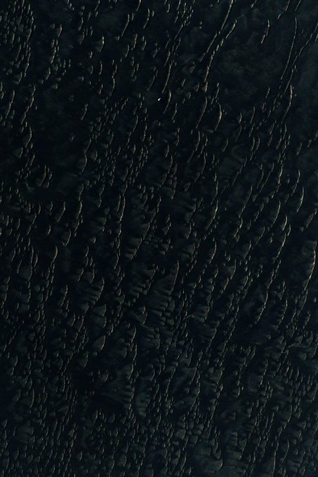 An aerial view of dark, undulating sand dunes captured in low light, where the rippled textures of the sand catch faint glimmers of illumination, evoking the "melting" quality referenced in the title. The abstract, textured composition mirrors the title "Melting Dunes III," translating the organic, fluid appearance of sand ripples into a generative artwork.