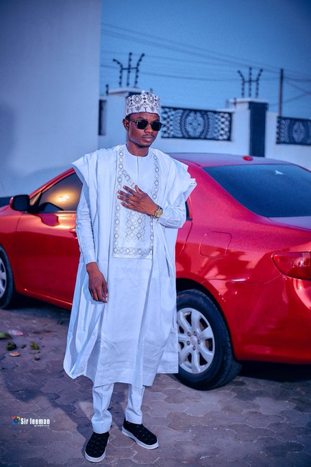 A man dressed in traditional white Nigerian attire with an embroidered cap and sunglasses poses in front of a red car, with a 'Sir leeman' watermark in the corner.