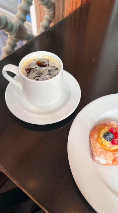A dark wooden table holds a white cup of black coffee on a saucer and a pastry topped with fruit on a white plate.