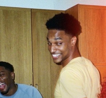 The image shows a smiling young man in a cream-colored shirt standing in front of wooden cabinets, with another person laughing in the background. The photo appears to be a candid, nostalgic-looking snapshot that complements the post's discussion of Ansem's financial success from PumpFun creator fees, though the image itself doesn't contain any visible text or numbers relating to the earnings mentioned.