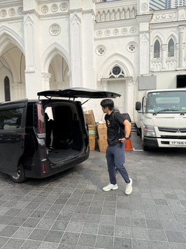 A man in a black shirt and grey pants stands next to the open trunk of a black van, appearing to unload items, with a white truck and ornate white building in the background.