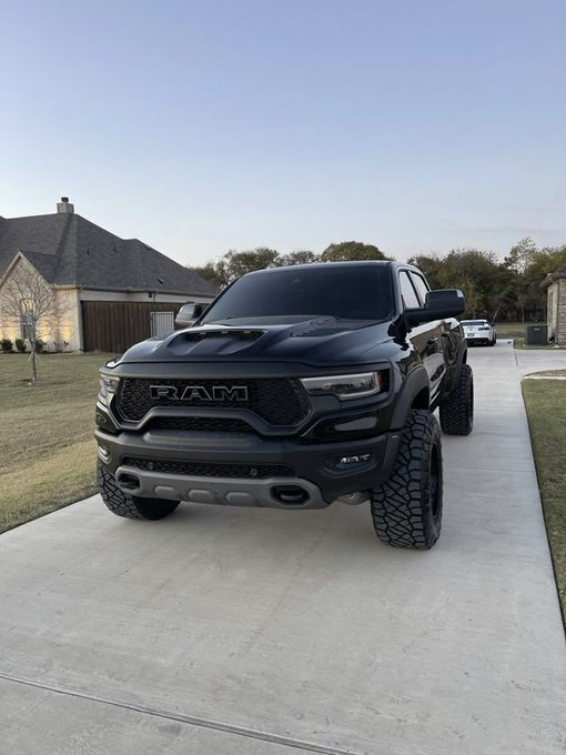 A matte black Ram TRX pickup truck with a hood scoop and large off-road tires sits parked on a concrete driveway in front of a suburban home, with the "RAM" badge clearly displayed on the grille, fitting the accompanying post that simply highlights the truck as "TRX 🔥."