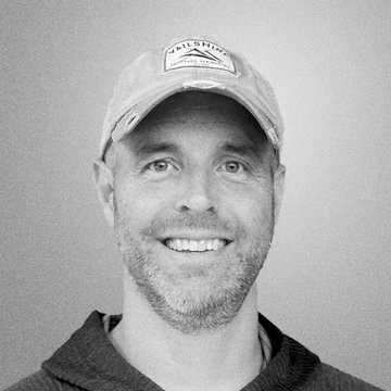 A black and white headshot portrait of a smiling man with a short beard wearing a cap with a patch that reads 'SAILRIG' and a hooded jacket.