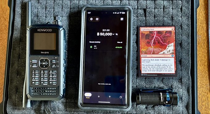 Top-down photo of items on a foam-lined case: a Kenwood TH-D75 handheld radio, a smartphone displaying a Robinhood portfolio with $50,000 and +$31.99, an Olight flashlight, and a Magic: The Gathering 'Lightning Bolt' card.