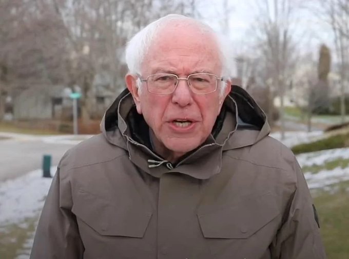 An older man with white hair and glasses, wearing a brown winter jacket, speaks outdoors in a snowy residential area.