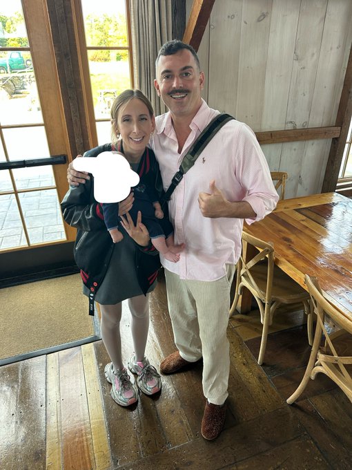 A smiling family of three—a woman, a man with a mustache giving a thumbs-up, and a baby in a carrier (face obscured by a white cloud)—posing indoors near a wooden table.