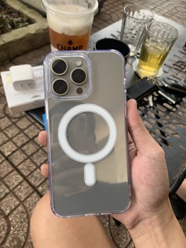 A hand holds a clear smartphone case with a white MagSafe ring, with an iced coffee drink and a glass of beer visible on a table in the background.
