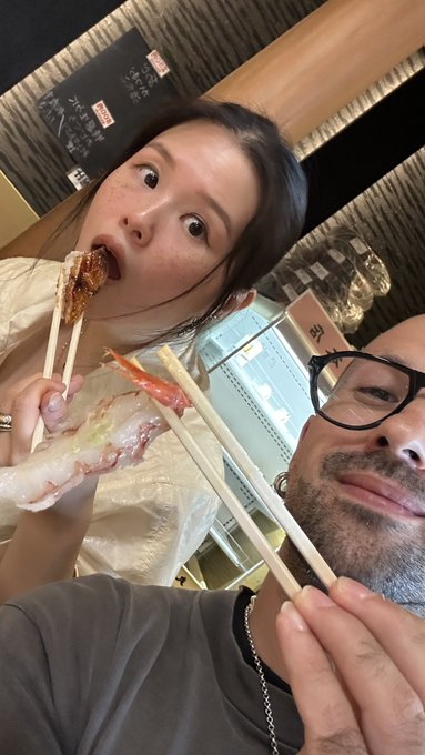 A woman takes a bite of sushi while a man holds up chopsticks beside her at a Japanese restaurant, matching the "gm japan maxxing" caption celebrating their morning in Japan. Japanese signage is visible on the wall in the background.