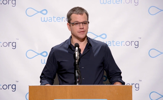 The image features a man standing at a podium with a microphone, giving a speech or presentation. The background of the image shows a banner that says "Water.org" and has a blue logo featuring a wave design. There are also other banners in the background, one of which displays the word "Matt Damon". The text on these banners suggests that there is an event related to water conservation or clean water initiatives taking place at this location.