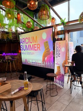 An Arcium "Summer Special Edition" event screen displaying colorful text and graphics, with attendees gathered around tables at what appears to be a meetup venue. The image visually captures the second meetup mentioned in the post, showcasing the Arcium event setup after the earlier XRPL Korea gathering.
