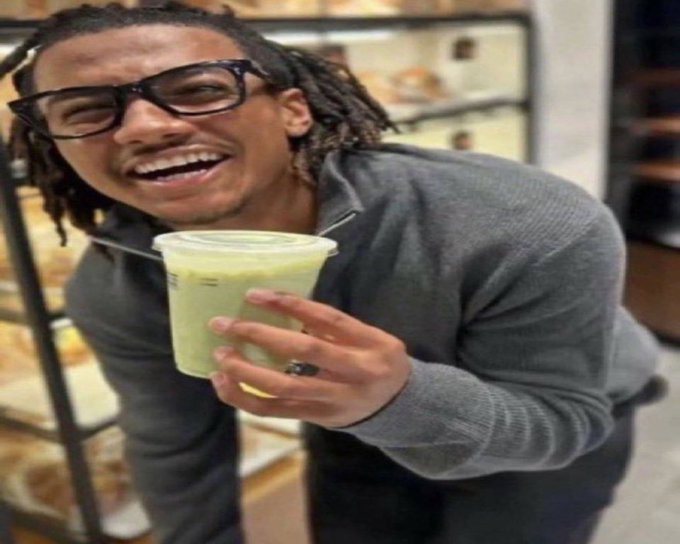 A person laughing while holding up a green drink, wearing glasses and a gray sweater. The image captures a triumphant, joyful moment that humorously conveys success after executing an exit strategy using @theunipcs for $JUGGERNAUT, embodying the feeling of waking up victorious after a successful trade.