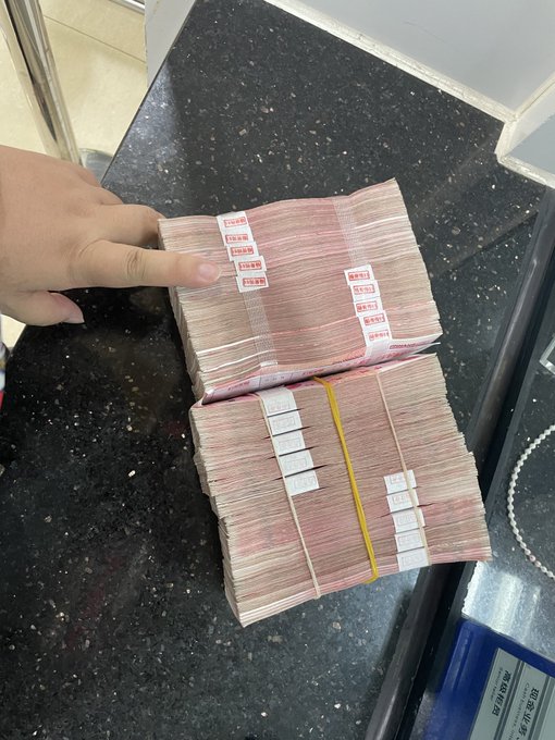 A hand rests on two thick bundles of pink Chinese currency (100 yuan notes) stacked on a dark granite countertop, with the bills banded together and labeled with price tags.