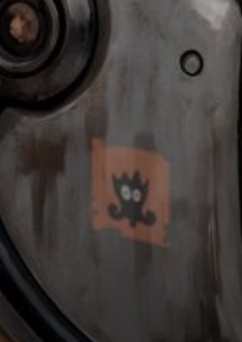 A close-up of a weathered, gray metallic surface with an orange rectangular sticker featuring a small black cartoonish creature logo.
