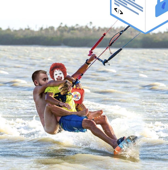 Two men kitesurfing on water embrace while holding the kite bar, with one wearing a Ronald McDonald mask and a yellow shirt with the McDonald's logo.