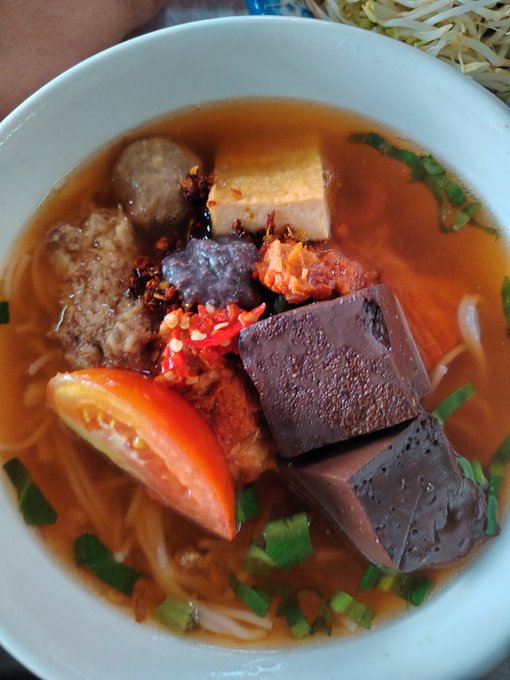A bowl of Vietnamese soup, likely bún riêu, containing noodles, tomato, tofu, meatballs, congealed blood cubes, and chili.