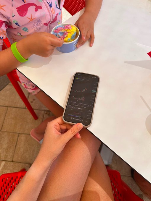 A child eating colorful ice cream at a table while another person holds a smartphone displaying a financial chart.