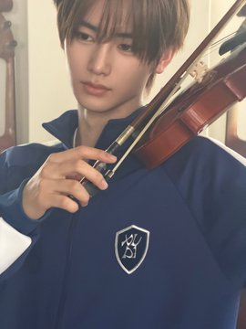 A young man with brown hair holds a violin near his face while wearing a navy blue jacket featuring a shield emblem with "K" and "1" inscribed on it. The image accompanies hashtags related to ALD1 and ANXIN, suggesting the person pictured may be associated with these groups or brands.