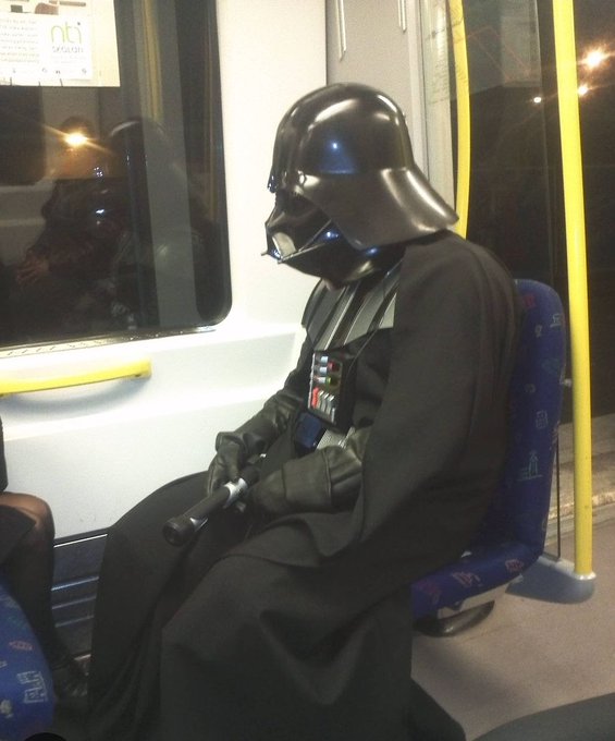 A person dressed in a Darth Vader costume, complete with helmet and lightsaber, sits on a public transit train. A sign in the window reads "nti" (partially visible, likely "INTI"), adding an ironic backdrop to the post's joke about the proper spelling of "lightsaber."