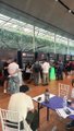 A blurry, low-resolution image showing several people gathered outdoors, possibly at a market or event, with tables and chairs visible in a covered space.
