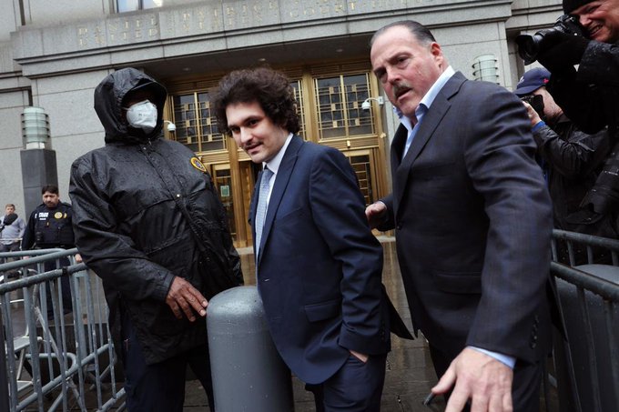 Former FTX CEO Sam Bankman-Fried in a navy suit walking past a masked individual outside a building, escorted by another man in a suit.